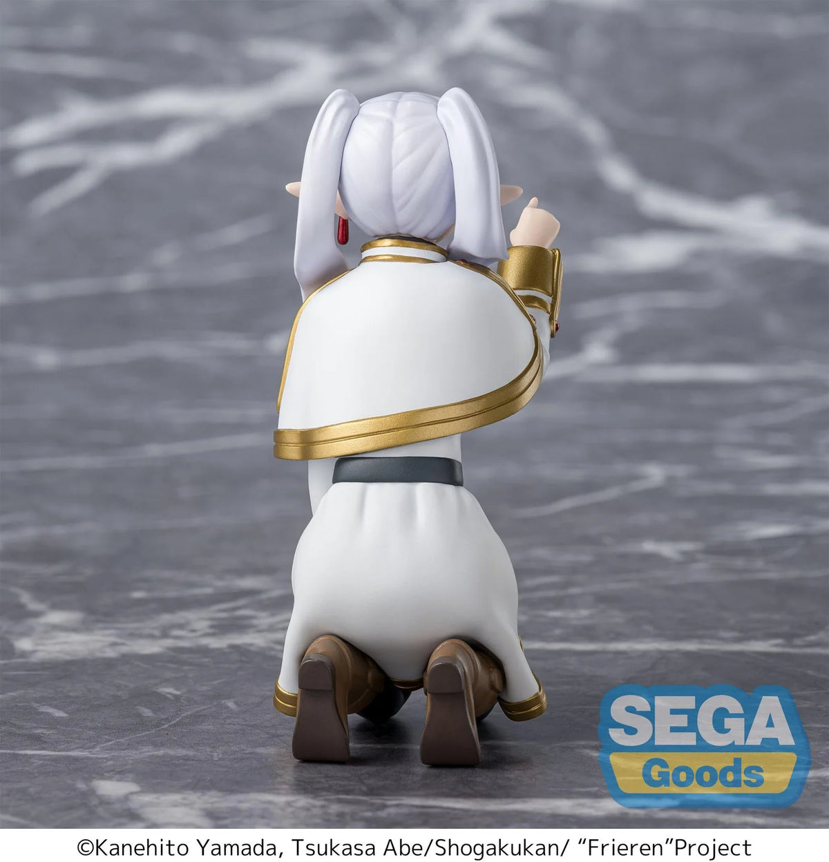 A back view of a Frieren: Beyond Journey's End SEGA Good figures character, showing the figure kneeling on a gray marble-textured surface. The figure has white hair in twin pigtails, a white robe with gold trim, and brown boots.