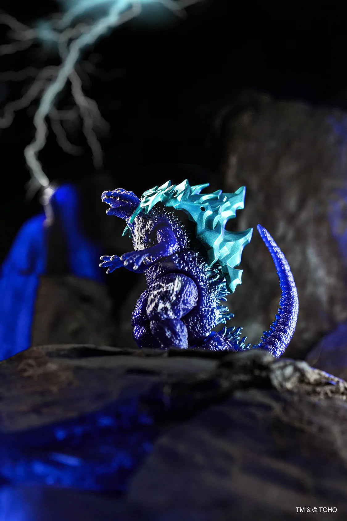 A blue and purple Godzilla action figure posed on a dark rocky surface with dramatic blue lighting and a blurred lightning background. - Action Figures - Movies - Godzilla & Kong