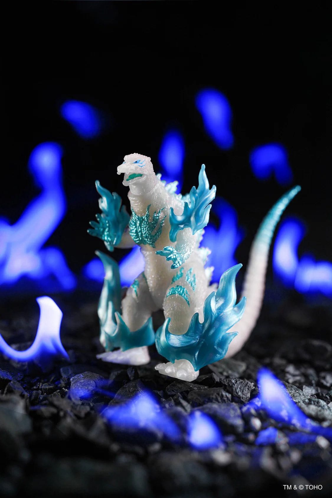 A small Godzilla figurine standing on dark pebbles with glowing blue flame-like effects in the background. The figure is primarily off-white with bright icy blue translucent flame-like accents around its body and tail.