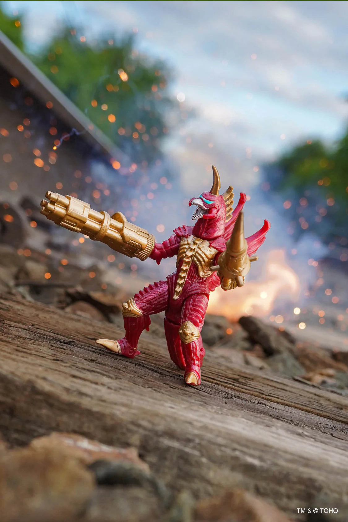 A red and gold Gigan action figure posed on a wooden surface with a blurred outdoor scene behind it, holding a large gold weapon. The background shows sparks, smoke, and trees. - Action Figures - Movies - Godzilla & Kong