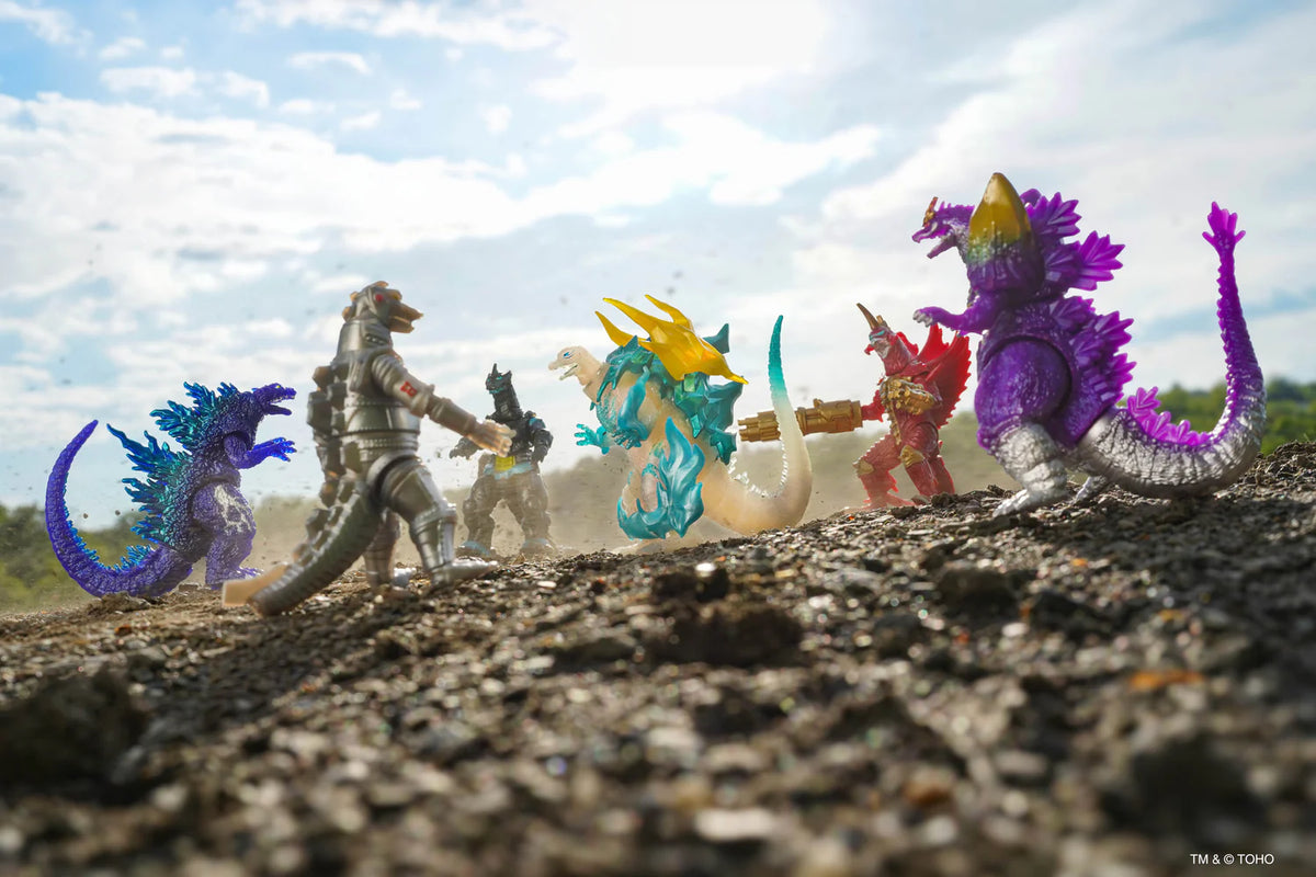 A group of colorful toy kaiju figures posed on a gravel surface outdoors under a cloudy sky. The figures include blue, purple, gray, teal, yellow, red, and white designs.
