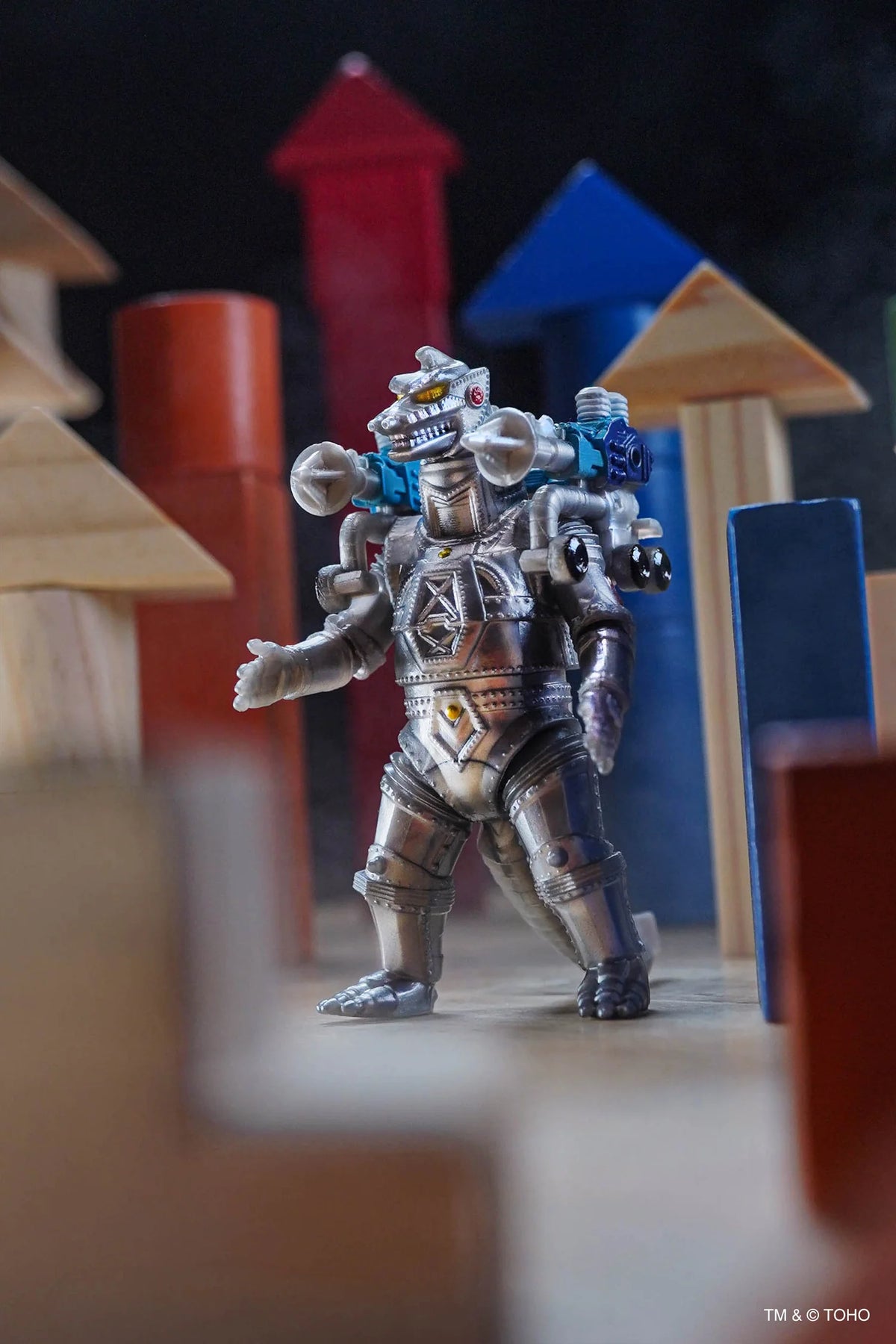 A small MechaGodzilla action figure stands in a toy-built scene with wooden blocks props. The figure appears silver and gray with small blue, white, and black details.