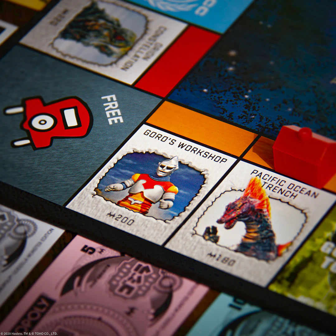 Close-up of a board game featuring Godzilla-themed property tiles, including 'Goro's Workshop' and 'Pacific Ocean Trench', with a red Hotel piece on a dark teal board.